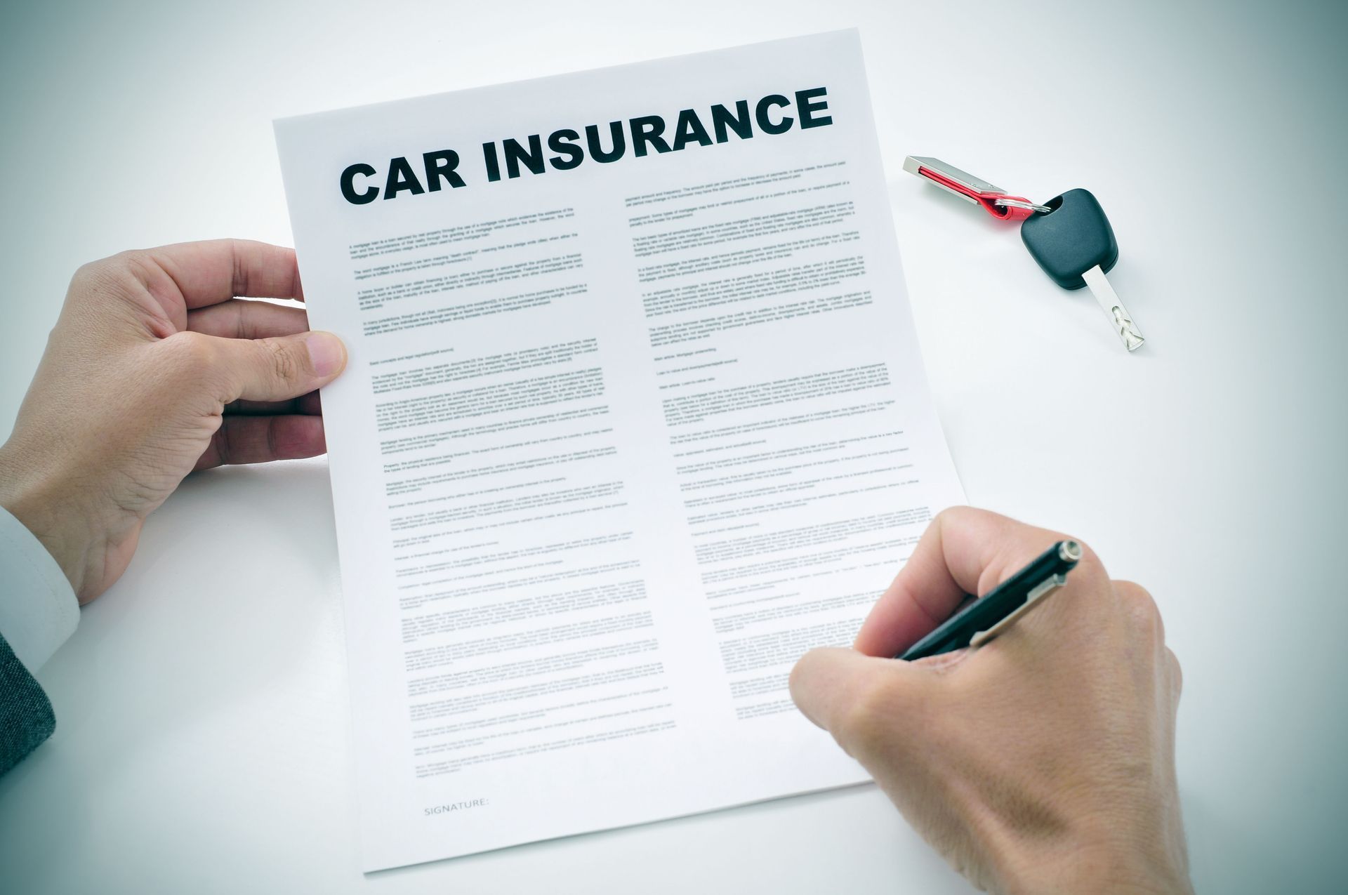 Hands holding a car insurance document, pen in hand, car keys nearby on a white surface.