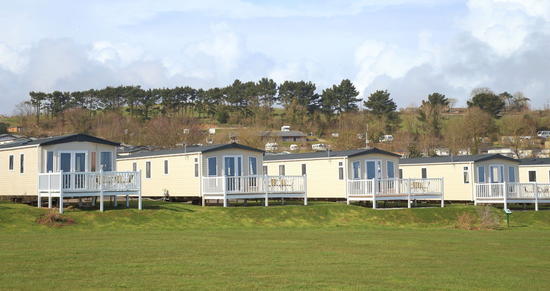 Row of white holiday cabins with small verandas in a field, with trees on a hillside in the background.