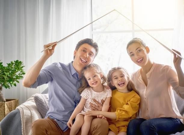 Family of four smiles under a roof symbol made by their arms, bright sunlight.