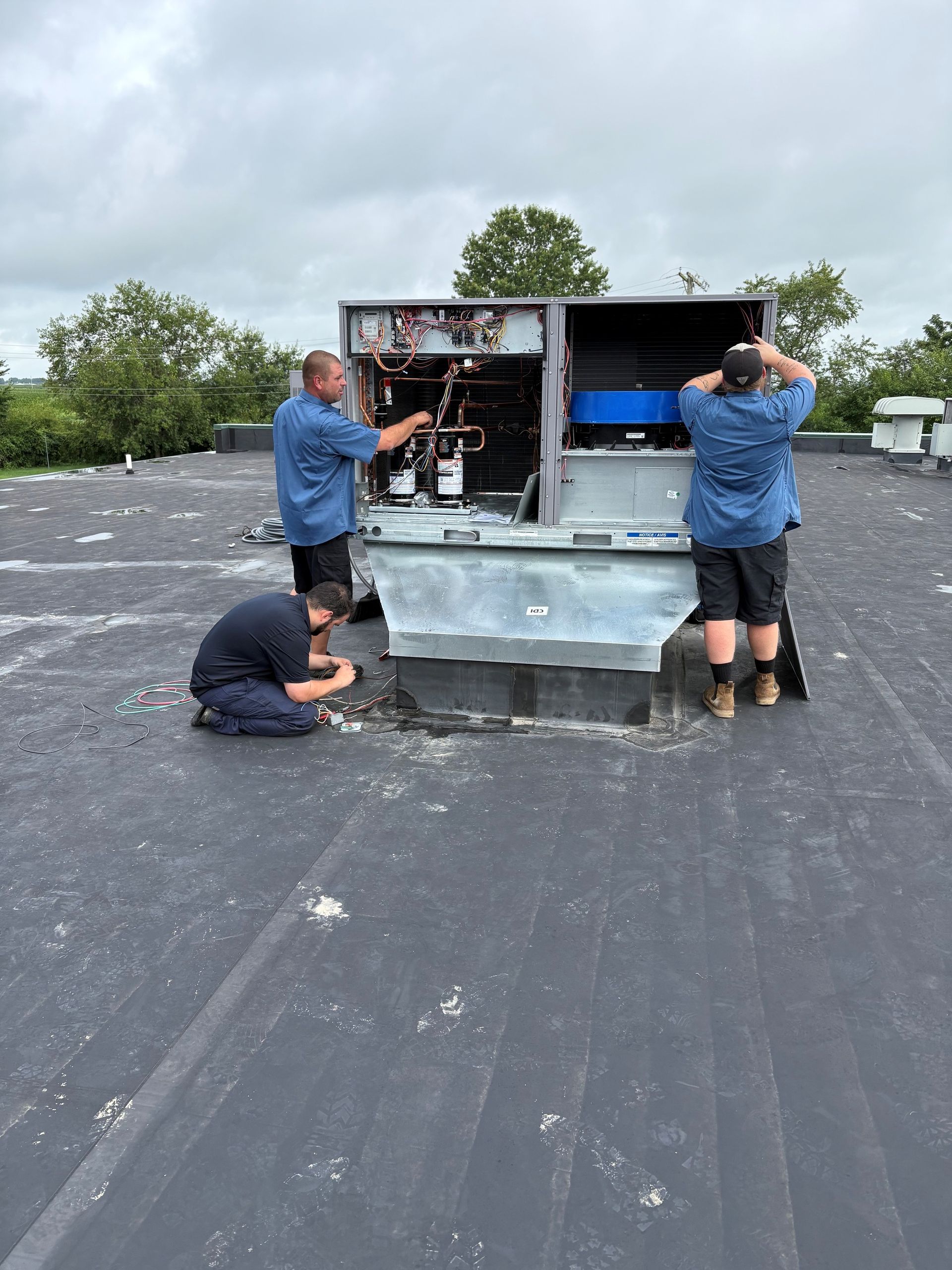 Three workers servicing rooftop HVAC units on a flat roof under cloudy skies