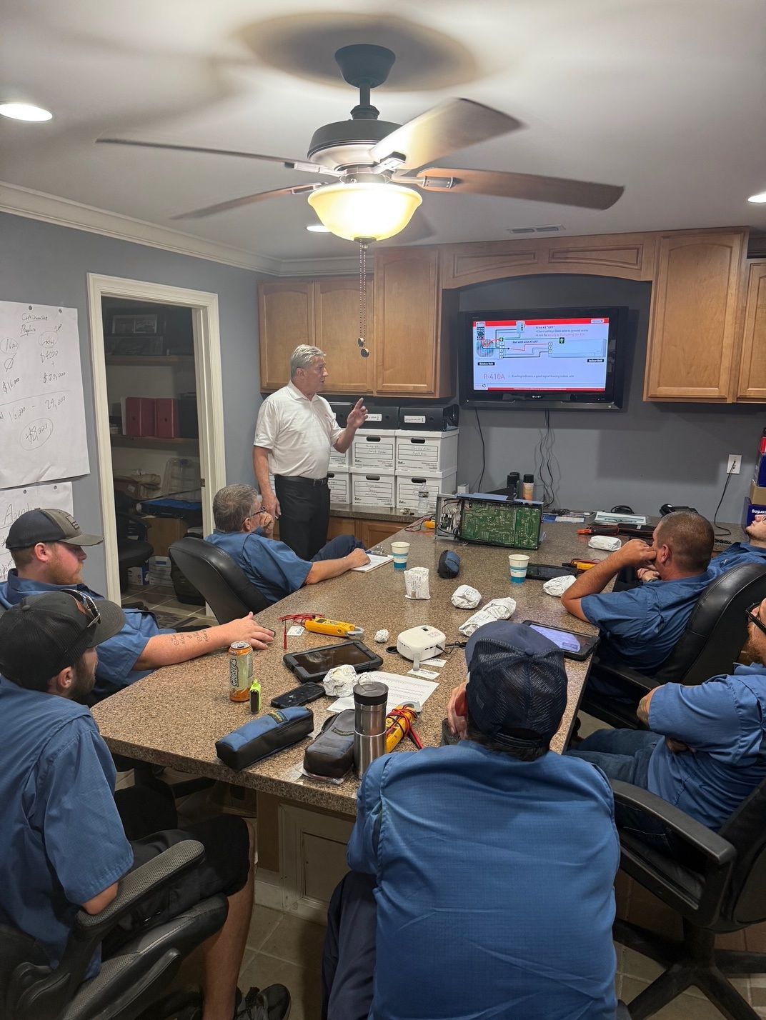 People gathered around a kitchen island watching a man present on a wall-mounted TV.