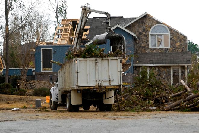A debris truck and excavator clearing storm damage beside a damaged house and fallen trees.