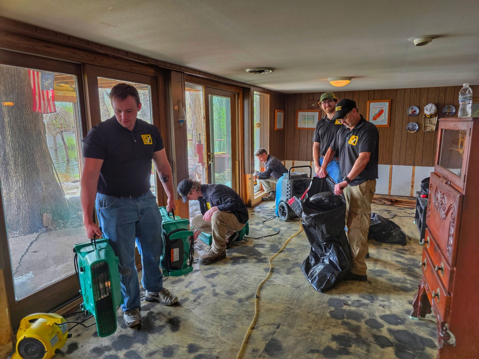 Workers cleaning water-damaged room with wet floor, open windows, and debris bags