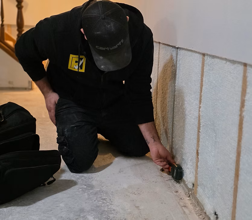 Person kneeling by a basement wall, inspecting a small pipe leak near the floor with a flashlight