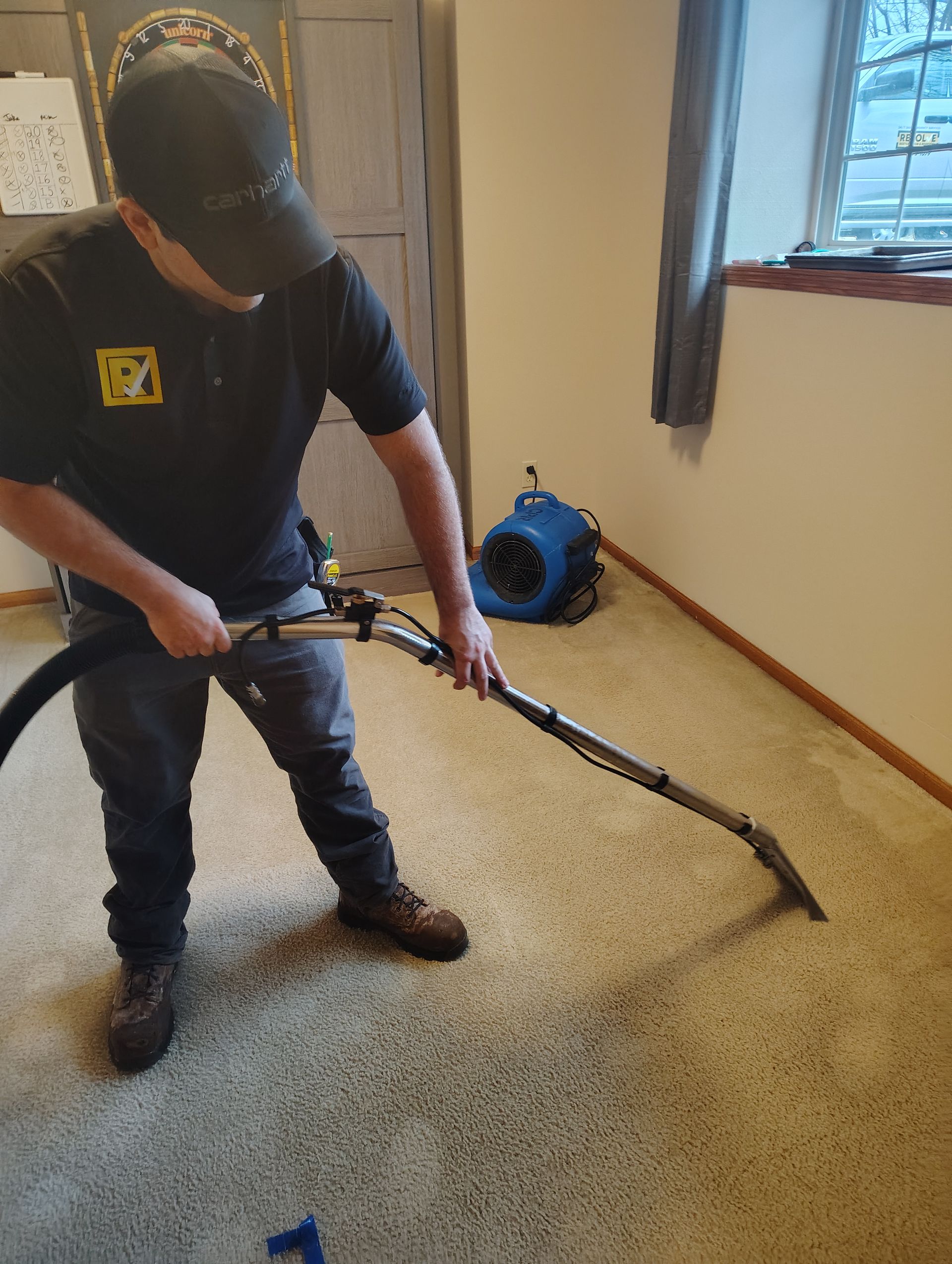 Person using a long-handled scraper on a carpeted floor near a window and blue fan