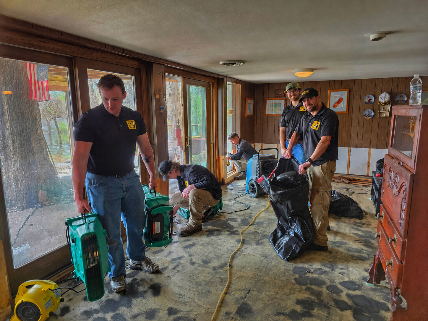 People cleaning a flooded room with wet floors, tools, and trash bags near windows and a doorway.