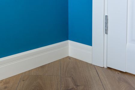 Corner of blue wall, white baseboard, and wood-look floor meets a white door frame with visible hinge.