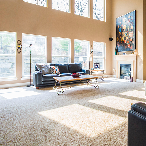 Spacious living room with large windows, blue sofa, and artwork over a fireplace. Beige walls and carpet.