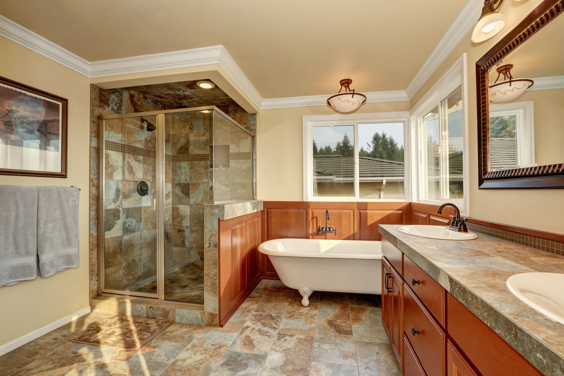 Bathroom with a claw-foot tub, stone shower, and brown cabinets. The walls are beige, and the floor is tiled.