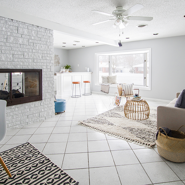 Living room with a white brick fireplace, white floors, and a rug. Sunlight streams in from a large window.