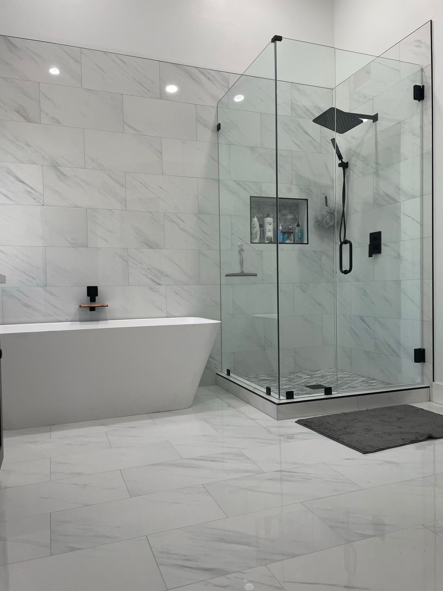 Modern bathroom with glass shower, white tub, and marble tile walls.