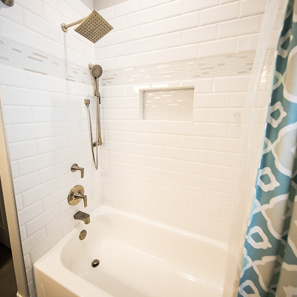White tiled shower with a handheld and rain shower head, built-in shelf, and blue patterned shower curtain.