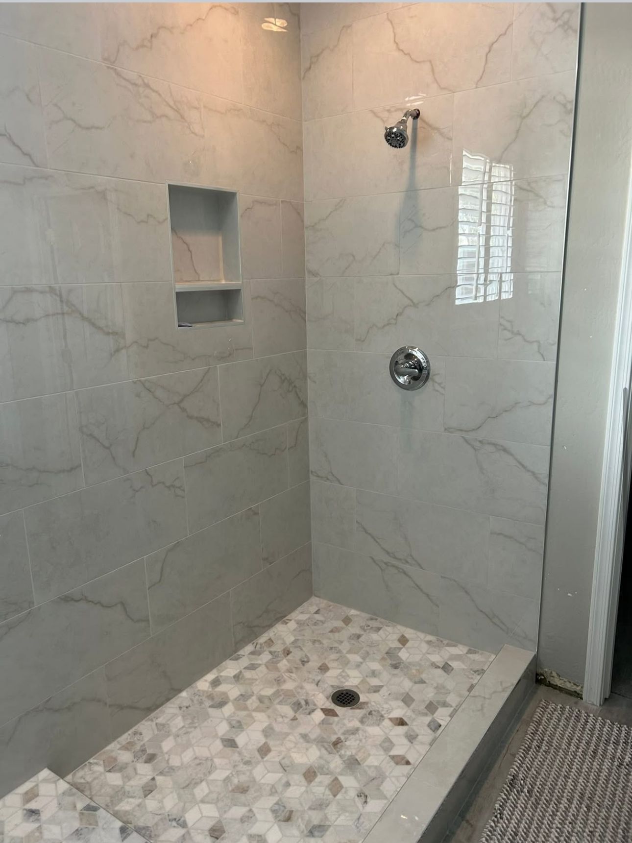 Bathroom shower with marble-like tile walls and a mosaic floor. Has a showerhead, control, and recessed shelf.