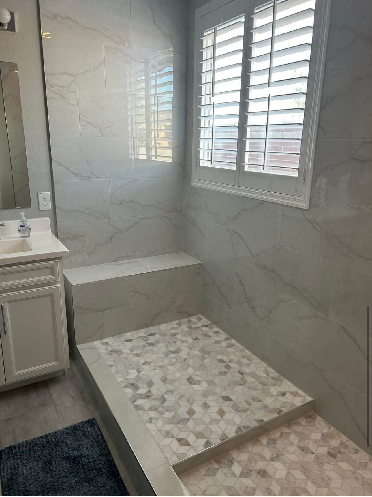 Bathroom with marble tile shower, built-in seat, and window with white shutters. White vanity.