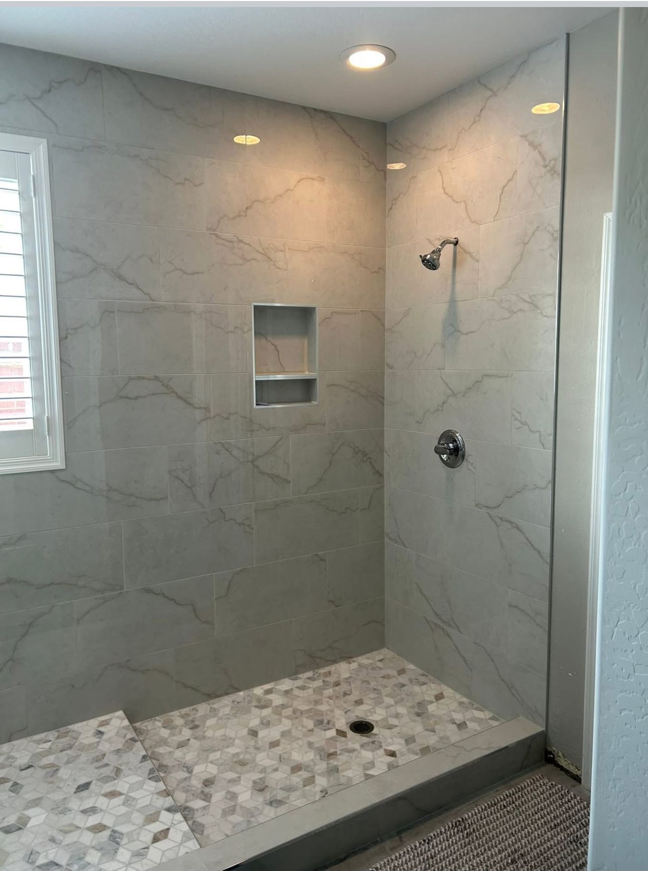 Tiled shower with glass panel, built-in shelf, and showerhead. Gray and white patterned tiles.