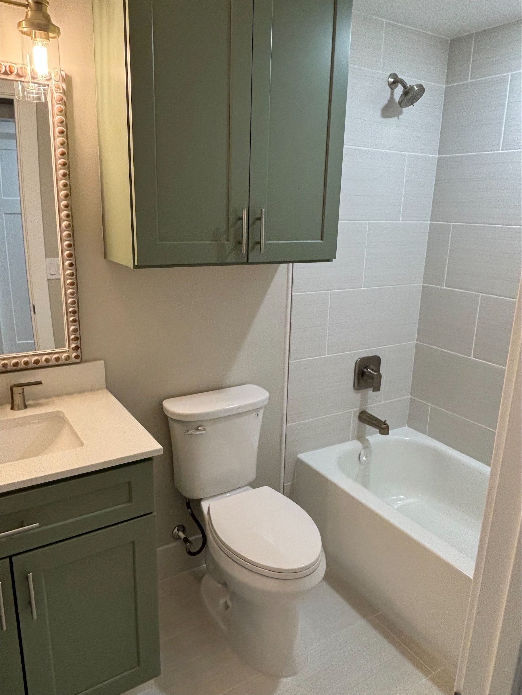 Bathroom with green cabinets, white toilet, bathtub, and gray tiled walls.