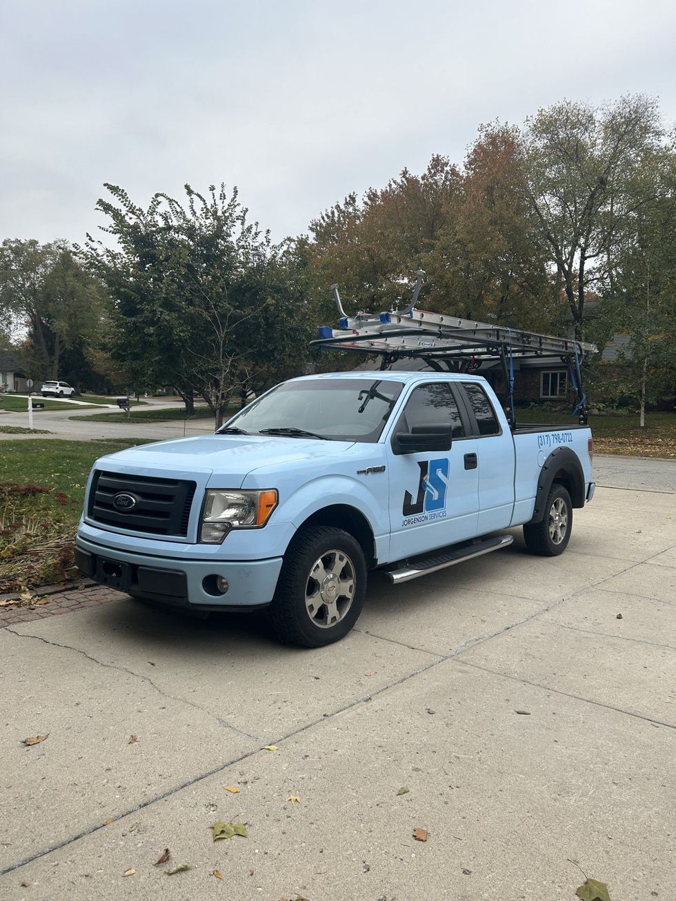 Light blue Ford pickup truck with ladder rack parked on pavement, logo on door.