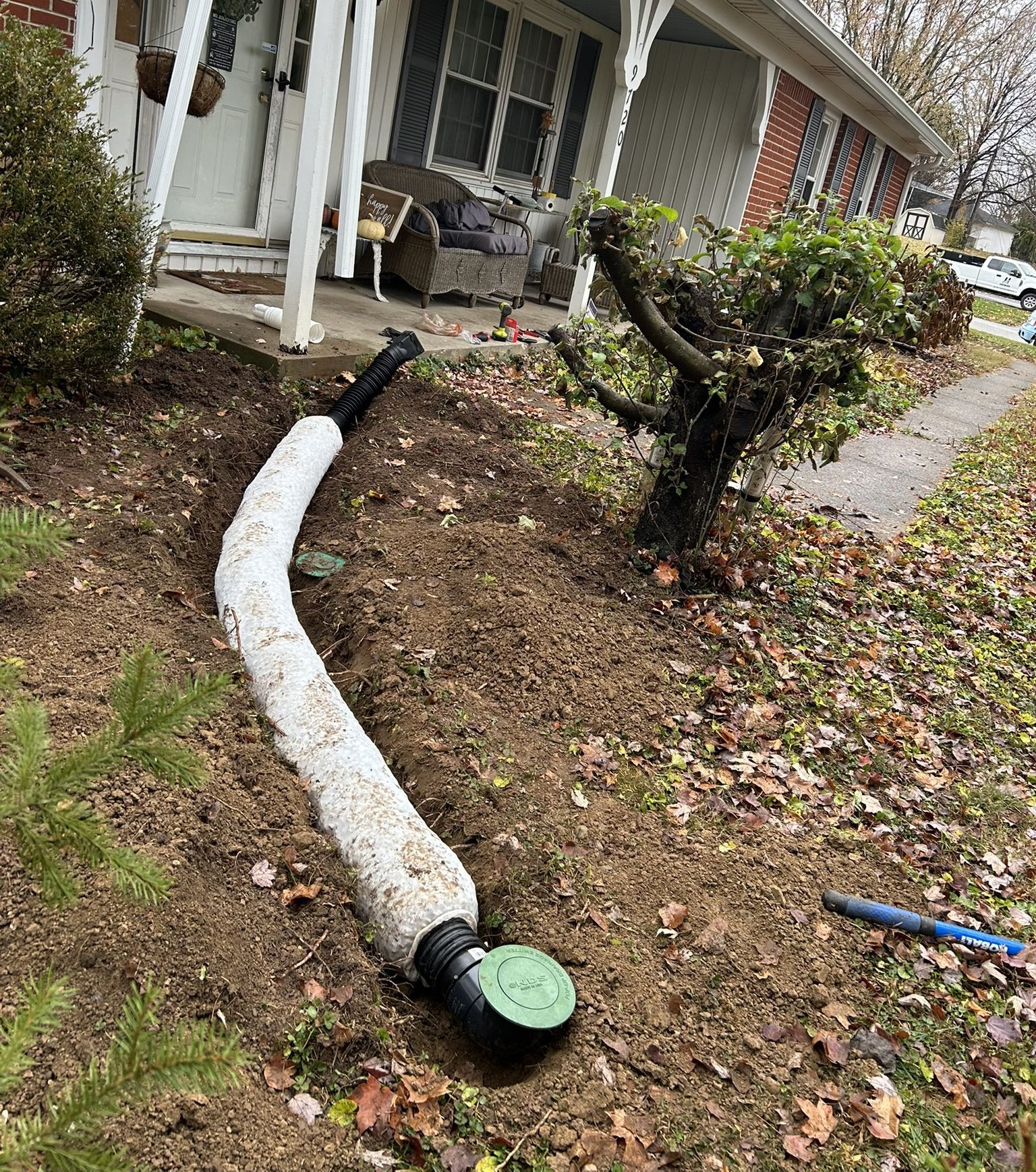 Drainage system installation in front yard: white corrugated pipe and green drain cover in dirt. House porch visible.