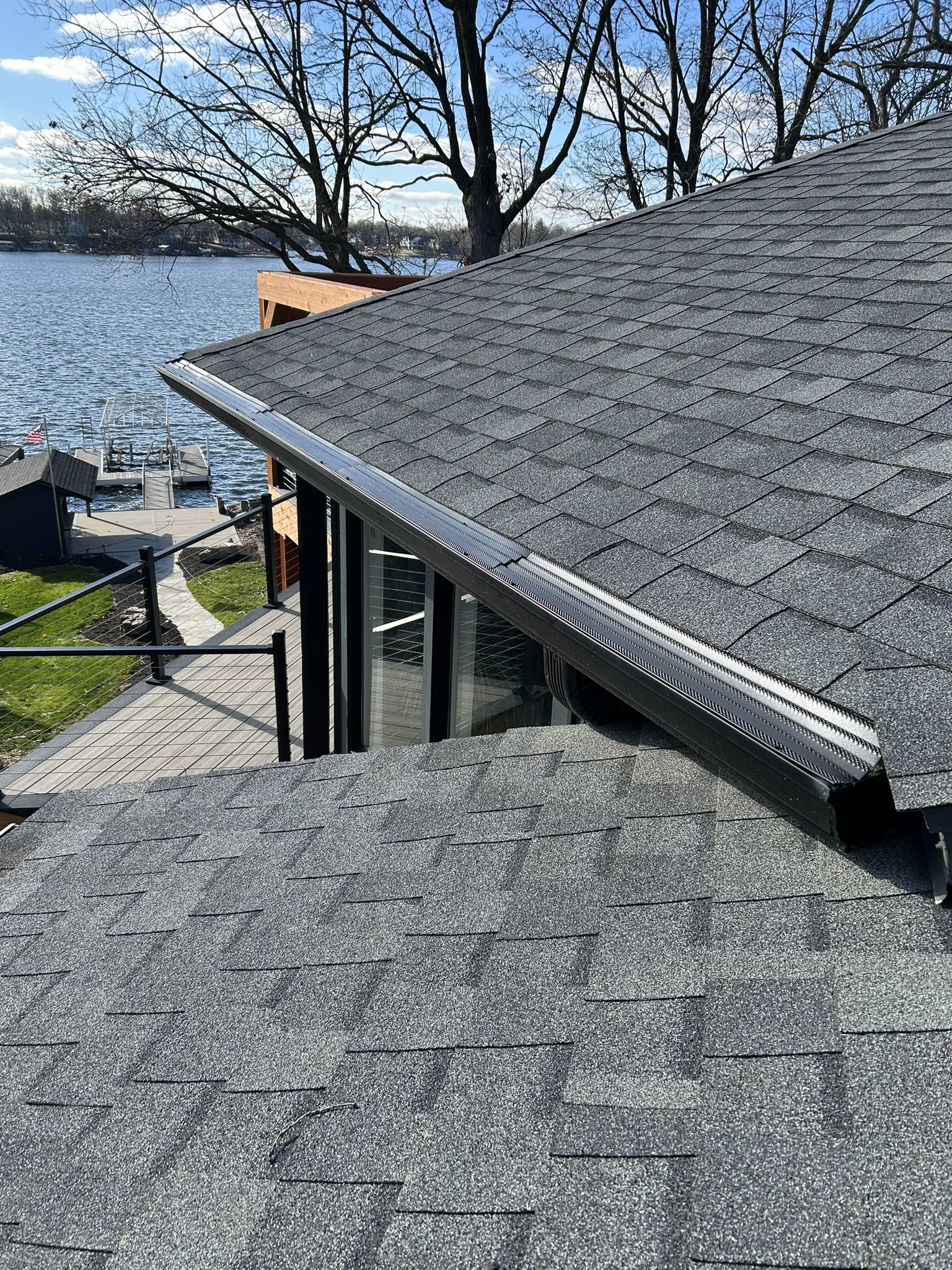 Close-up of a gray shingled roof with a black metal edge. Trees and water in the background.