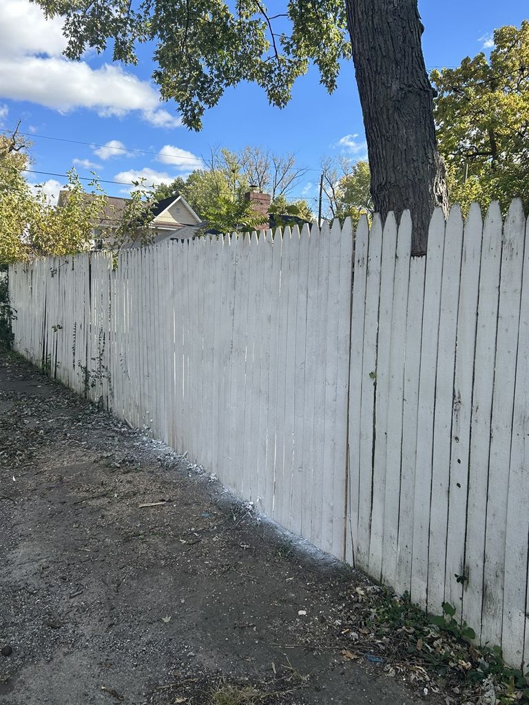 White picket fence along a gravel path, with a tree and houses in the background under a blue sky.