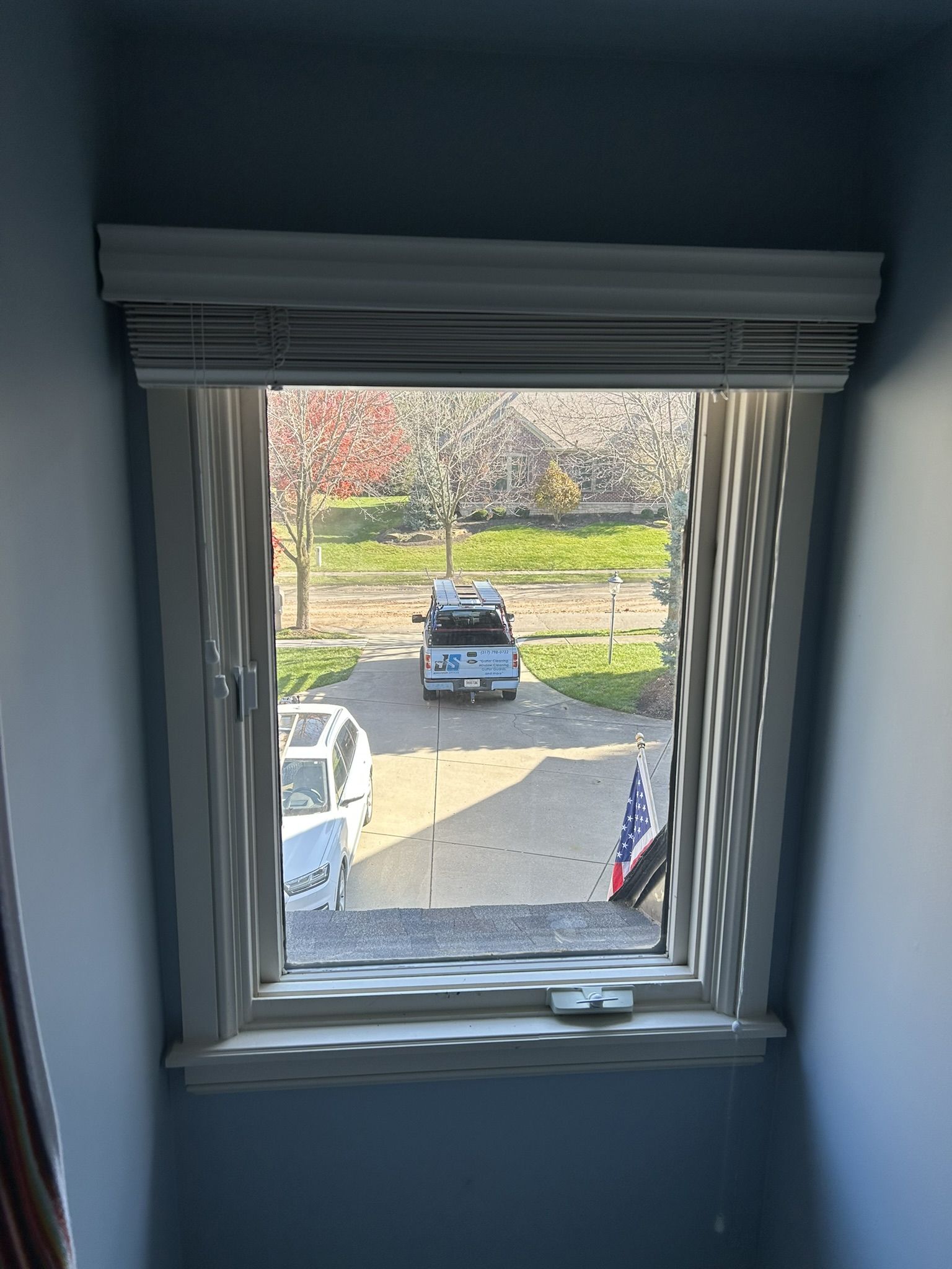 Window with blinds, looking out onto a driveway with vehicles, trees, and a flag. Sunlight casts shadows on the wall.