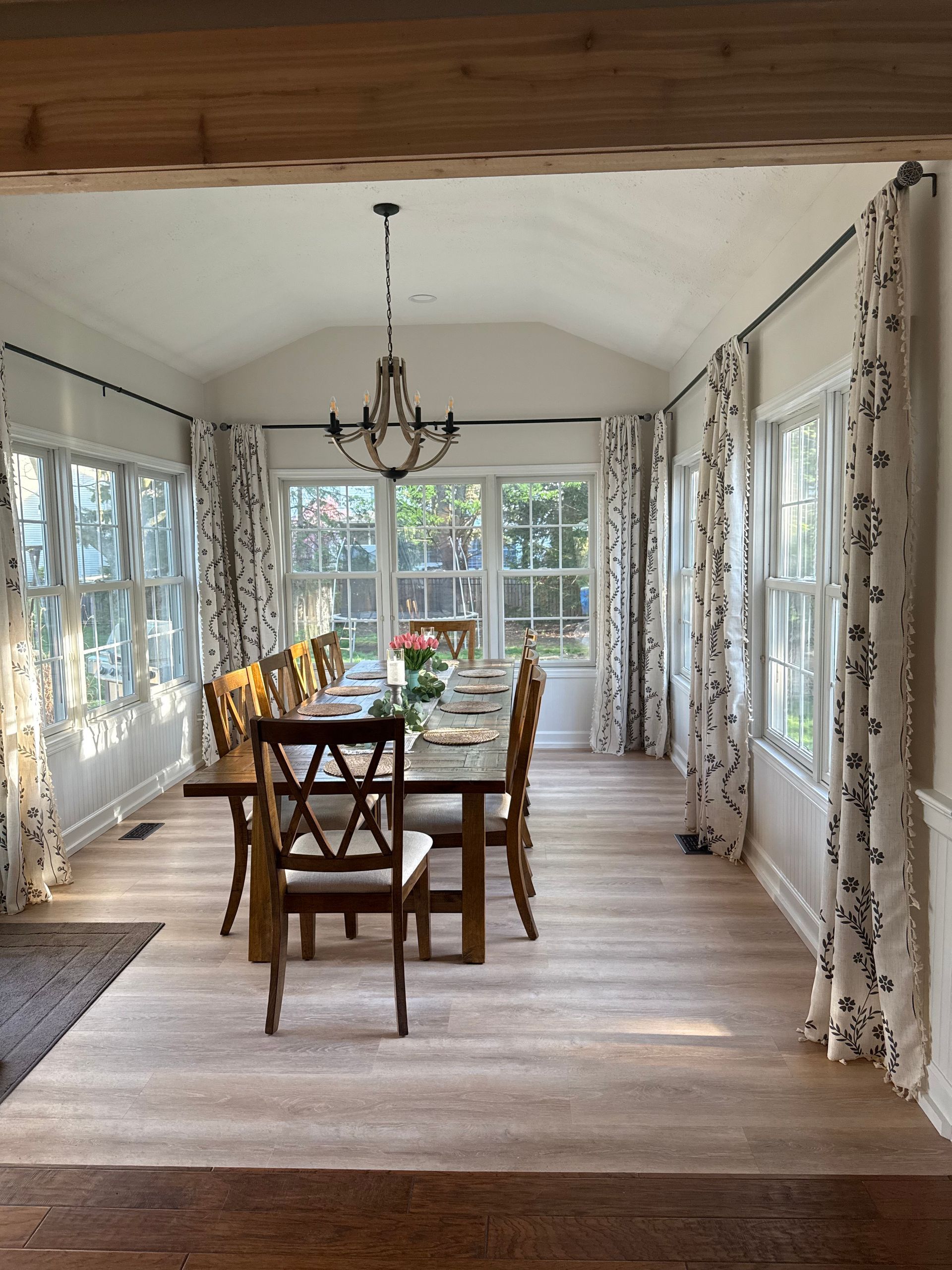 Dining room with a long wooden table, chairs, chandelier, and windows with patterned curtains.