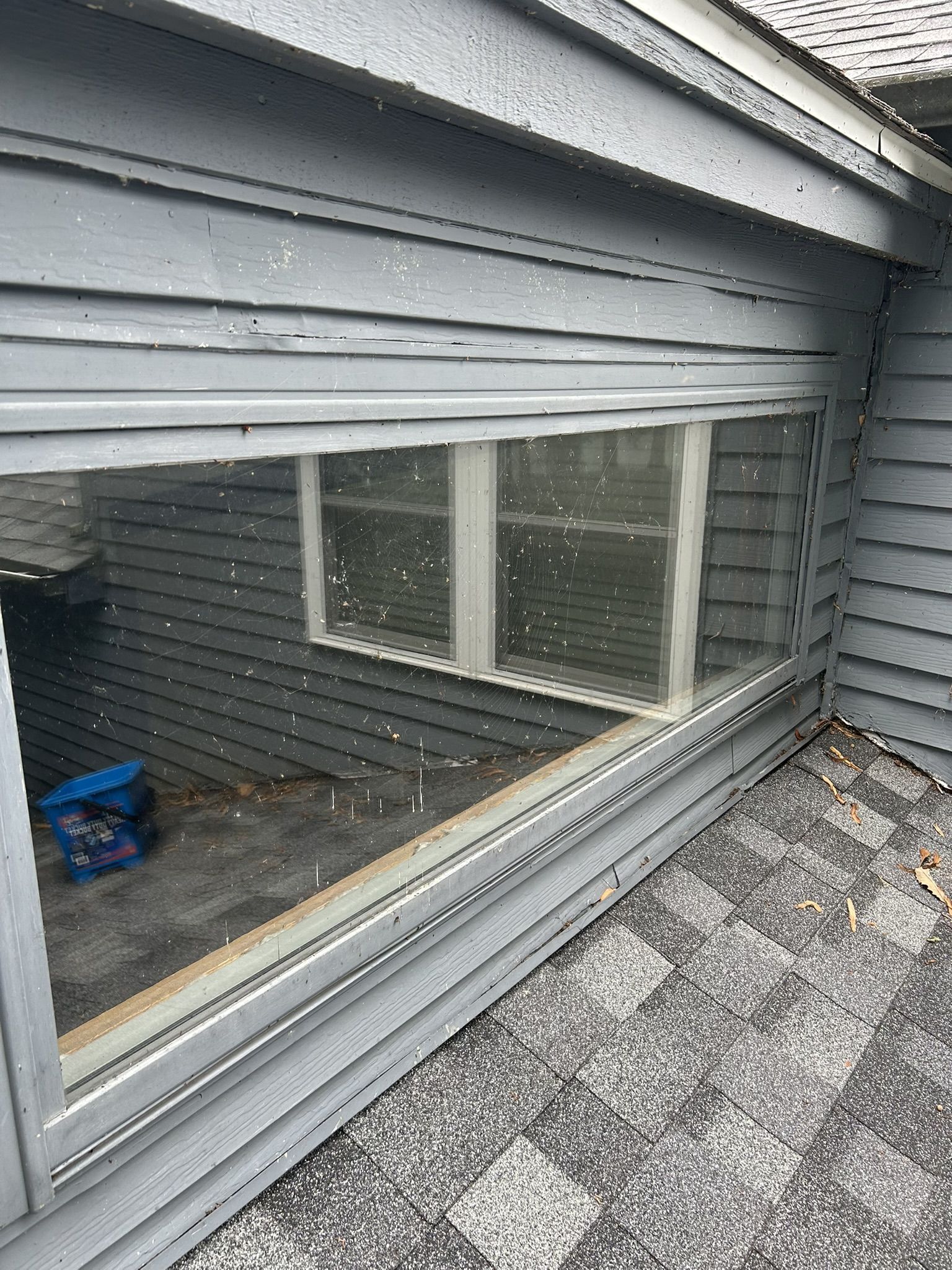 Exterior corner of a building with a large window. Gray siding, and a rooftop visible.