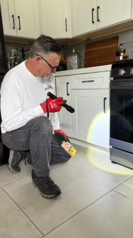 A person in work clothes kneels in a kitchen, using a flashlight and a yellow trap to inspect under the oven.