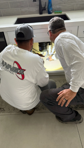 Two individuals in work attire crouch under a kitchen sink to inspect the plumbing and garbage disposal area.