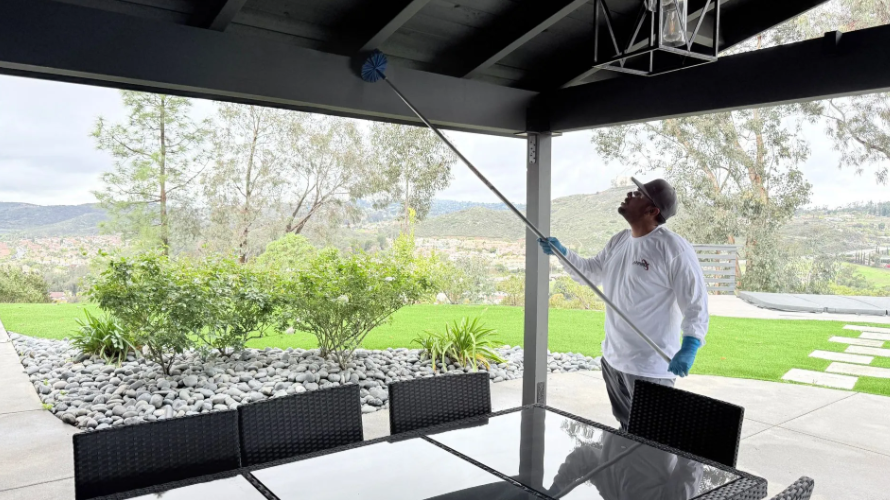 A person uses an extension pole to clean the underside of a dark patio roof with a view of a grassy yard in the background.