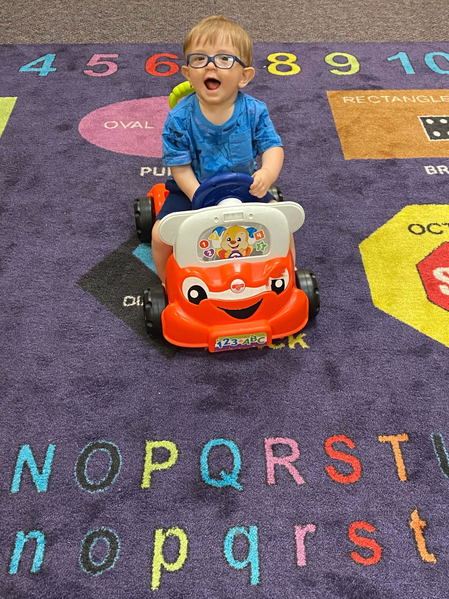A young boy is sitting on a toy car on a carpet.