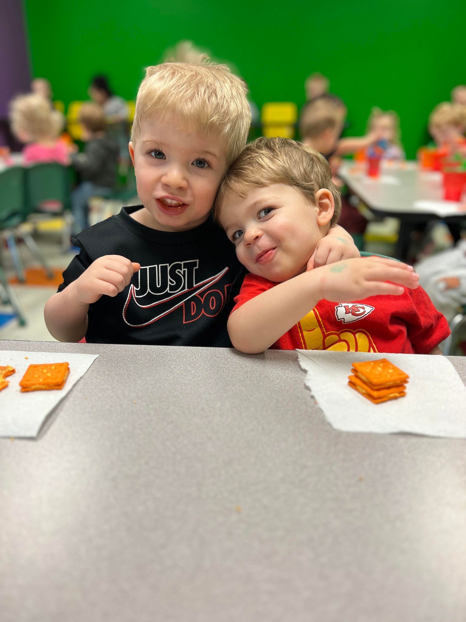 Two young boys are sitting at a table hugging each other.