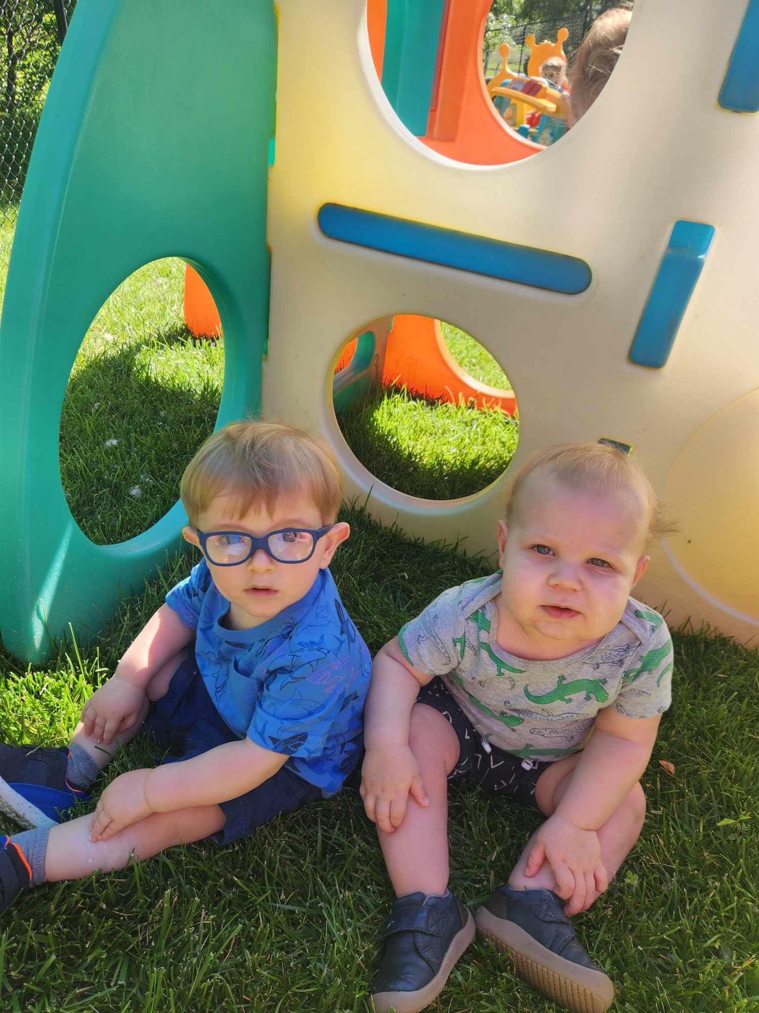 Two young boys are sitting on the grass in front of a playground.