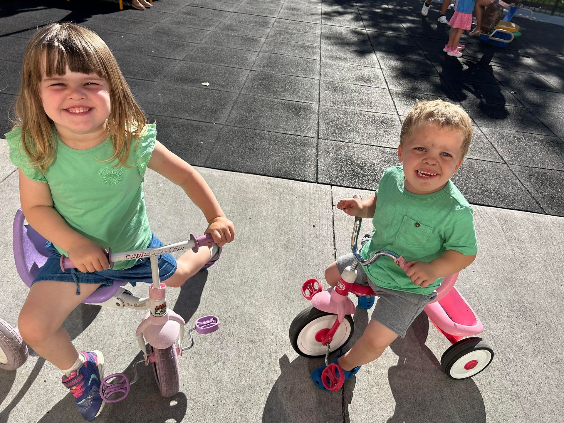 A boy and a girl are riding tricycles on a sidewalk.