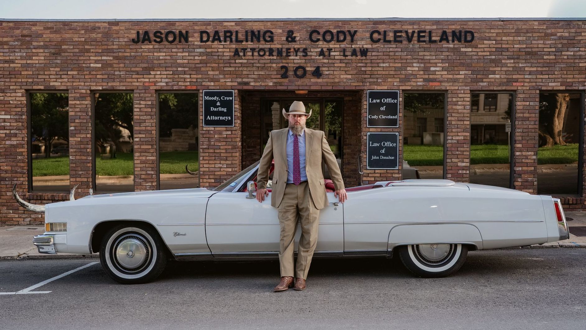 Man in a suit stands by a white convertible in front of a brick building