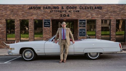 A man in a suit and tie is standing next to a white car in front of a cody cleveland building
