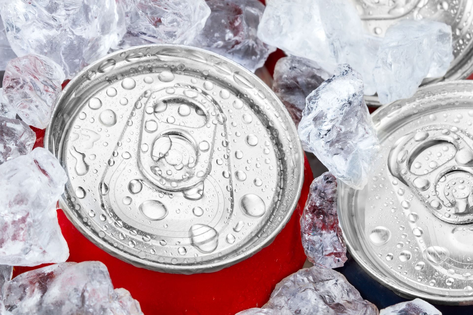 Close-up of two cold, silver soda cans with condensation, surrounded by ice cubes on a red surface.