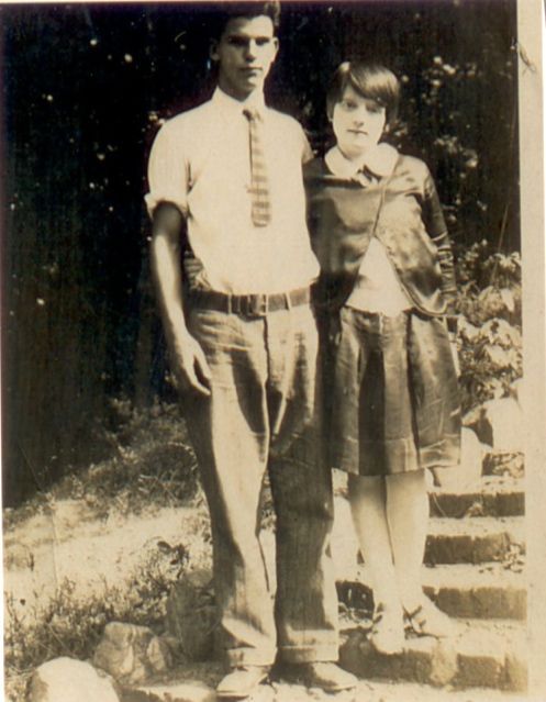 A young man and woman pose outdoors. The man wears a tie and rolled-up sleeves. The woman wears a pleated skirt.