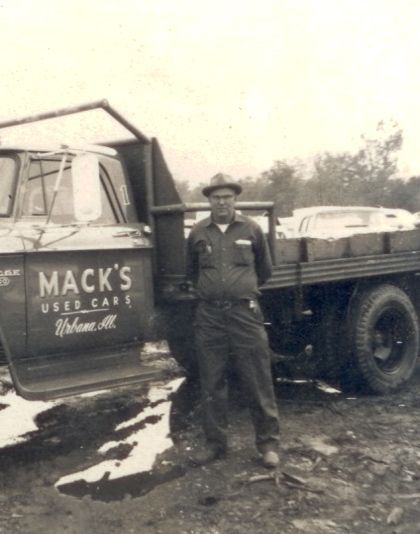 Man standing by Mack's Used Cars truck; the truck bed is full of items.