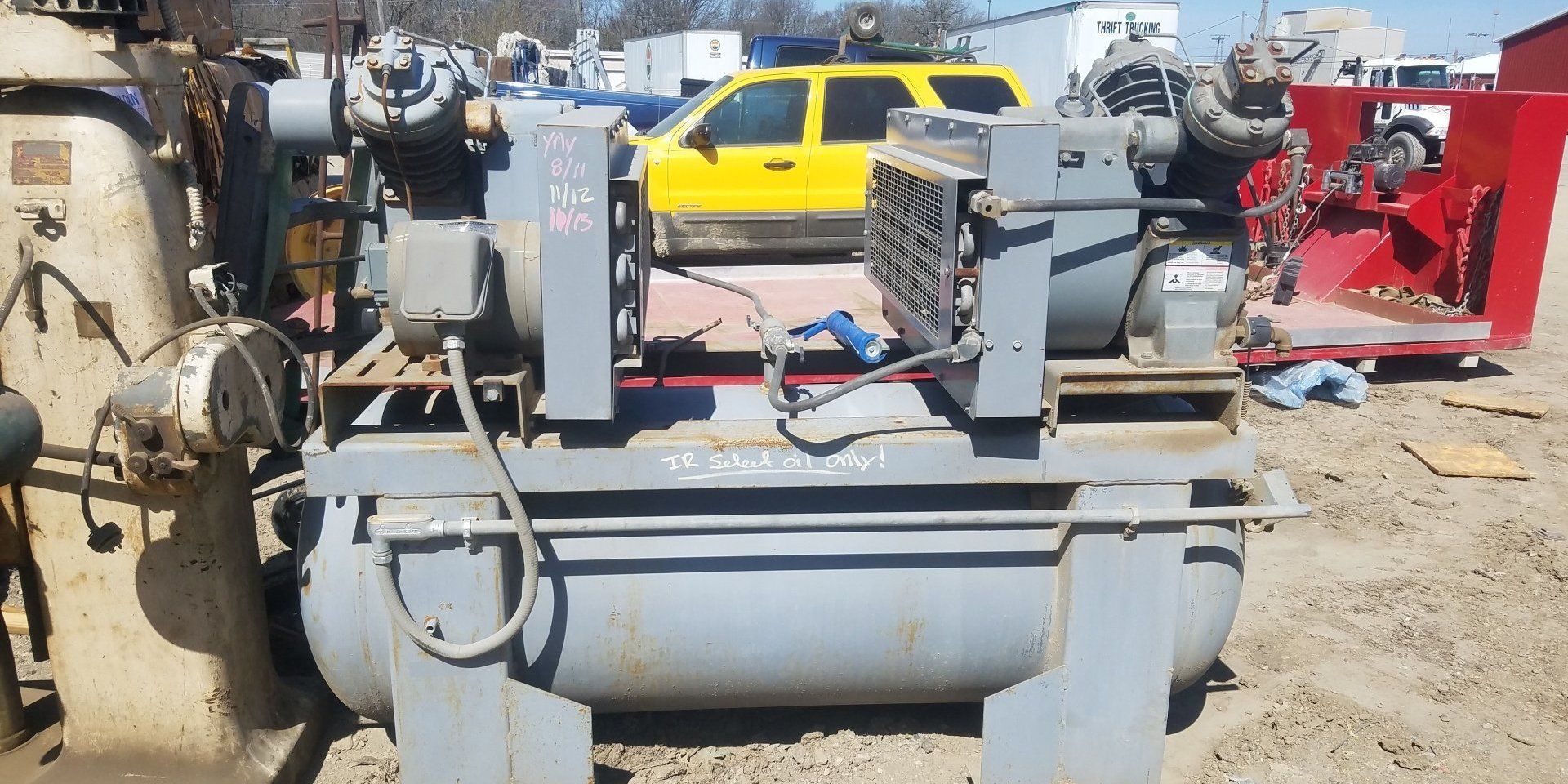 An industrial air compressor with tanks, in an outdoor setting, with a yellow SUV in the background.