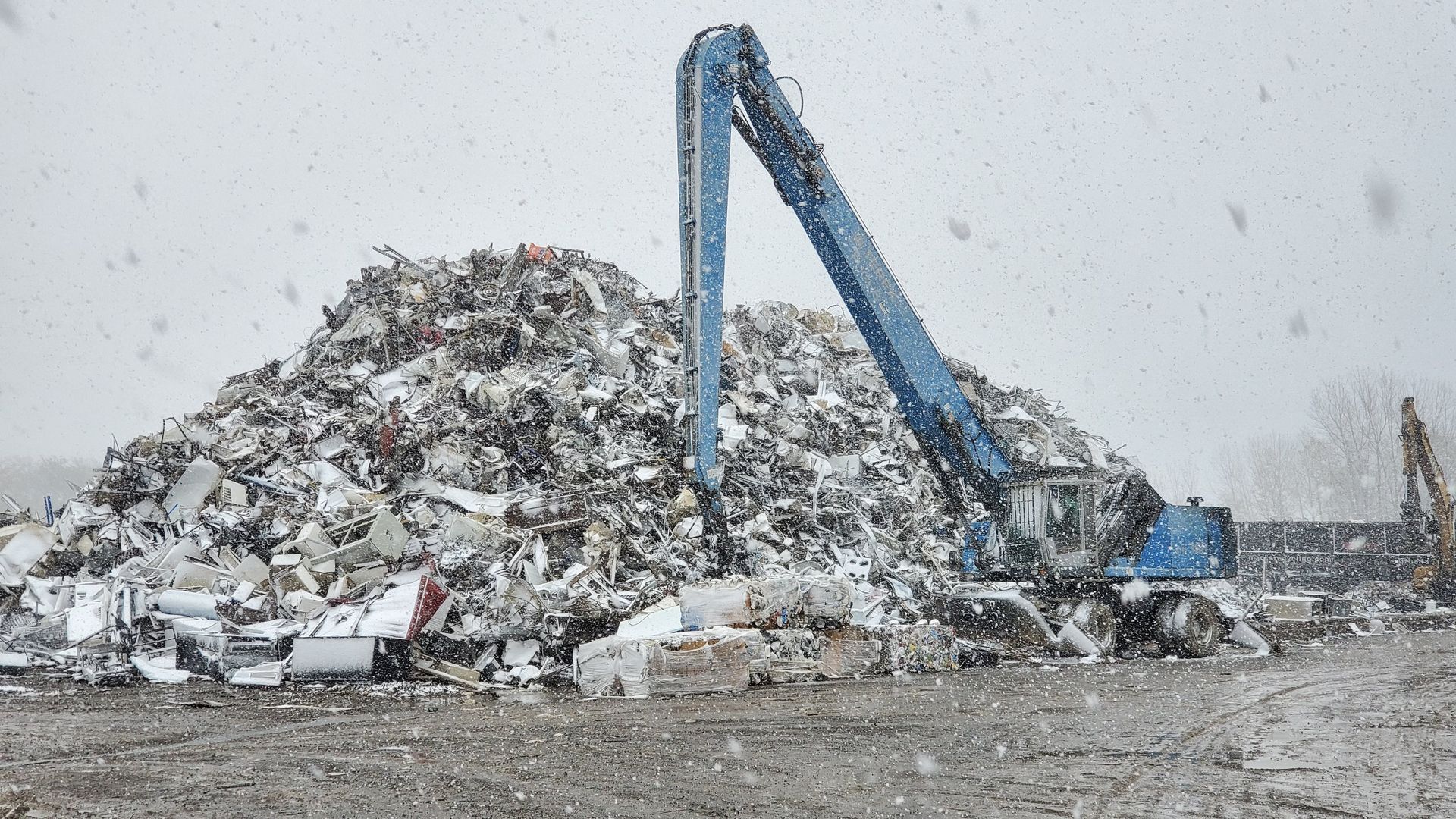 Blue excavator sorting scrap metal in a snowy outdoor junkyard.