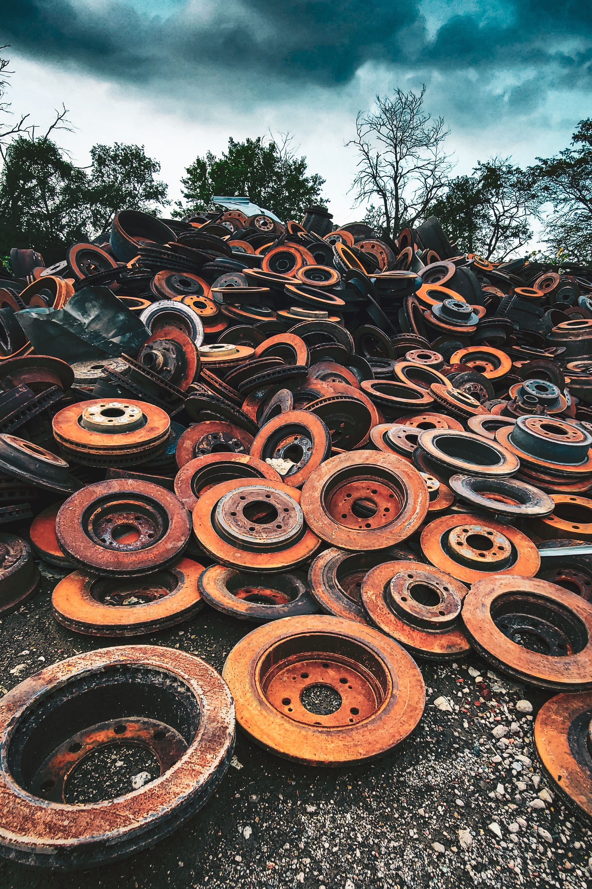 Pile of rusted car brake rotors; overcast sky and trees in background.