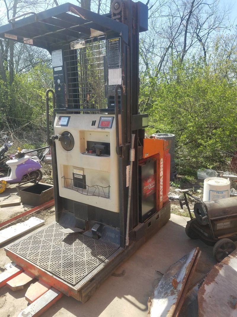 Stand-up forklift, beige and orange, outdoors. Metal platform, controls, and lifting mechanism.