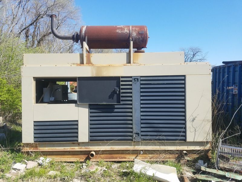 A weathered beige generator with a rusty exhaust pipe on a metal base outdoors.