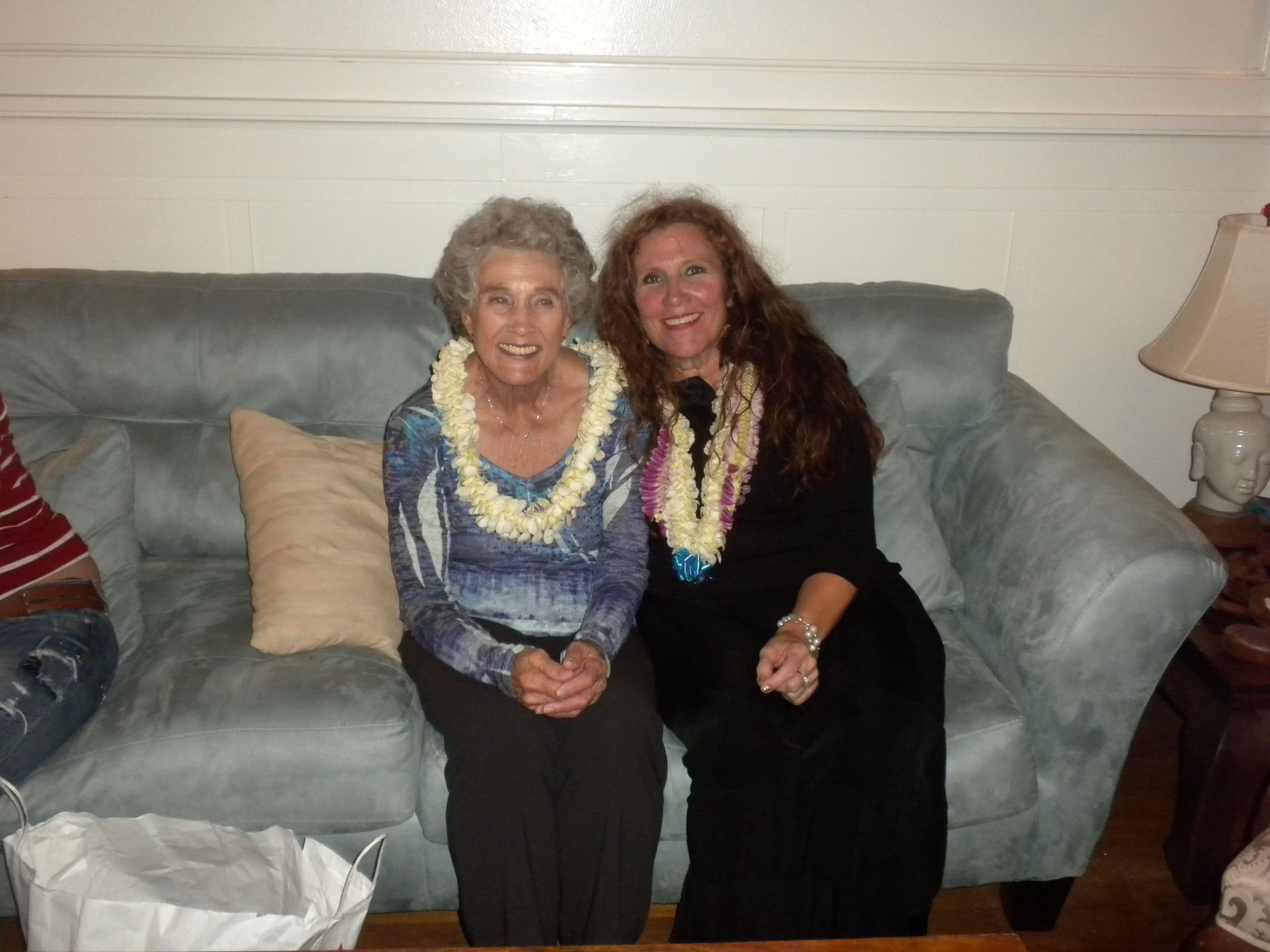 Two women in leis sit on a blue couch; one is older, smiling.