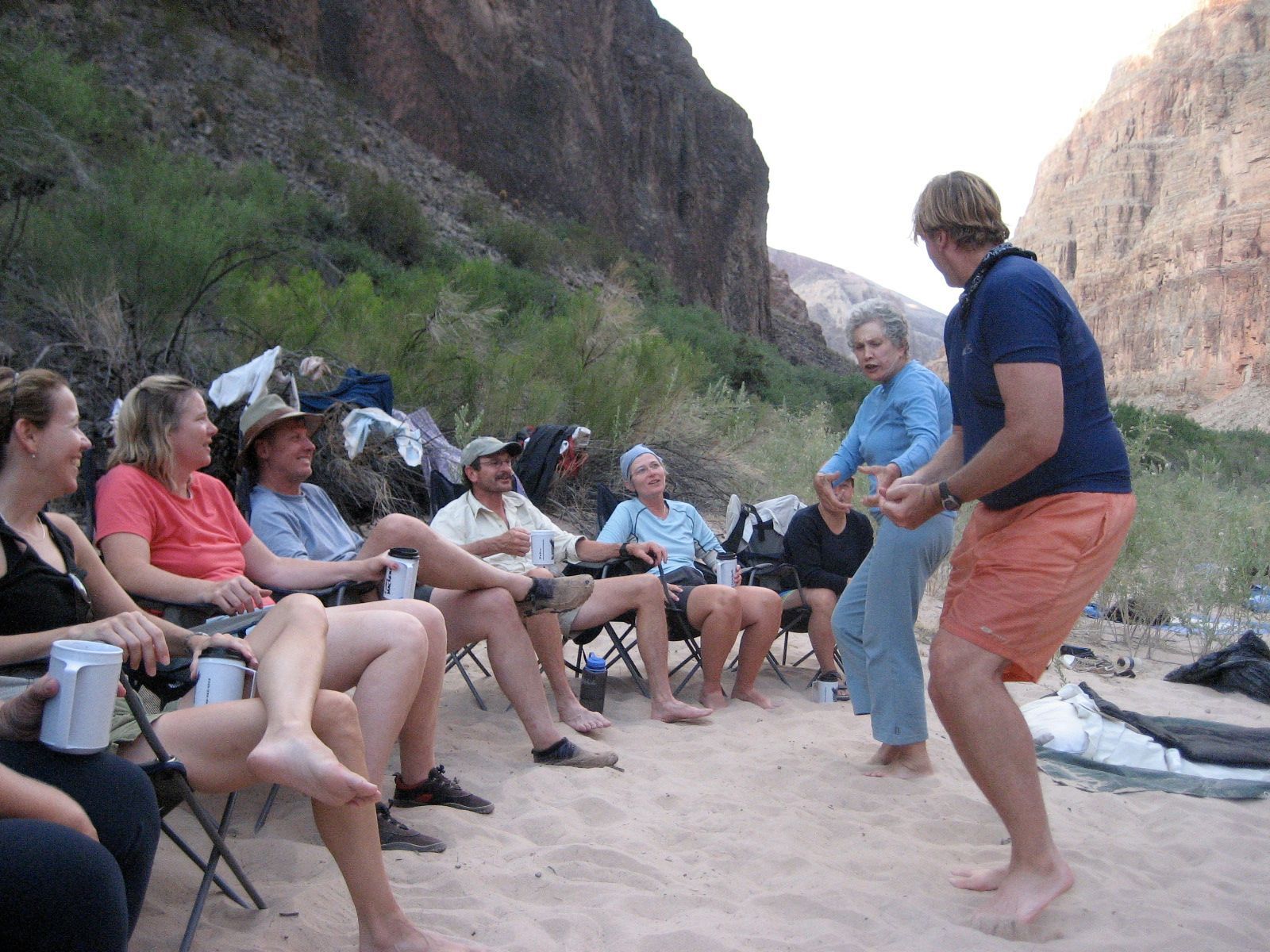 Group of people on a sandy riverbank, two people interacting in the center, canyon walls in the background.