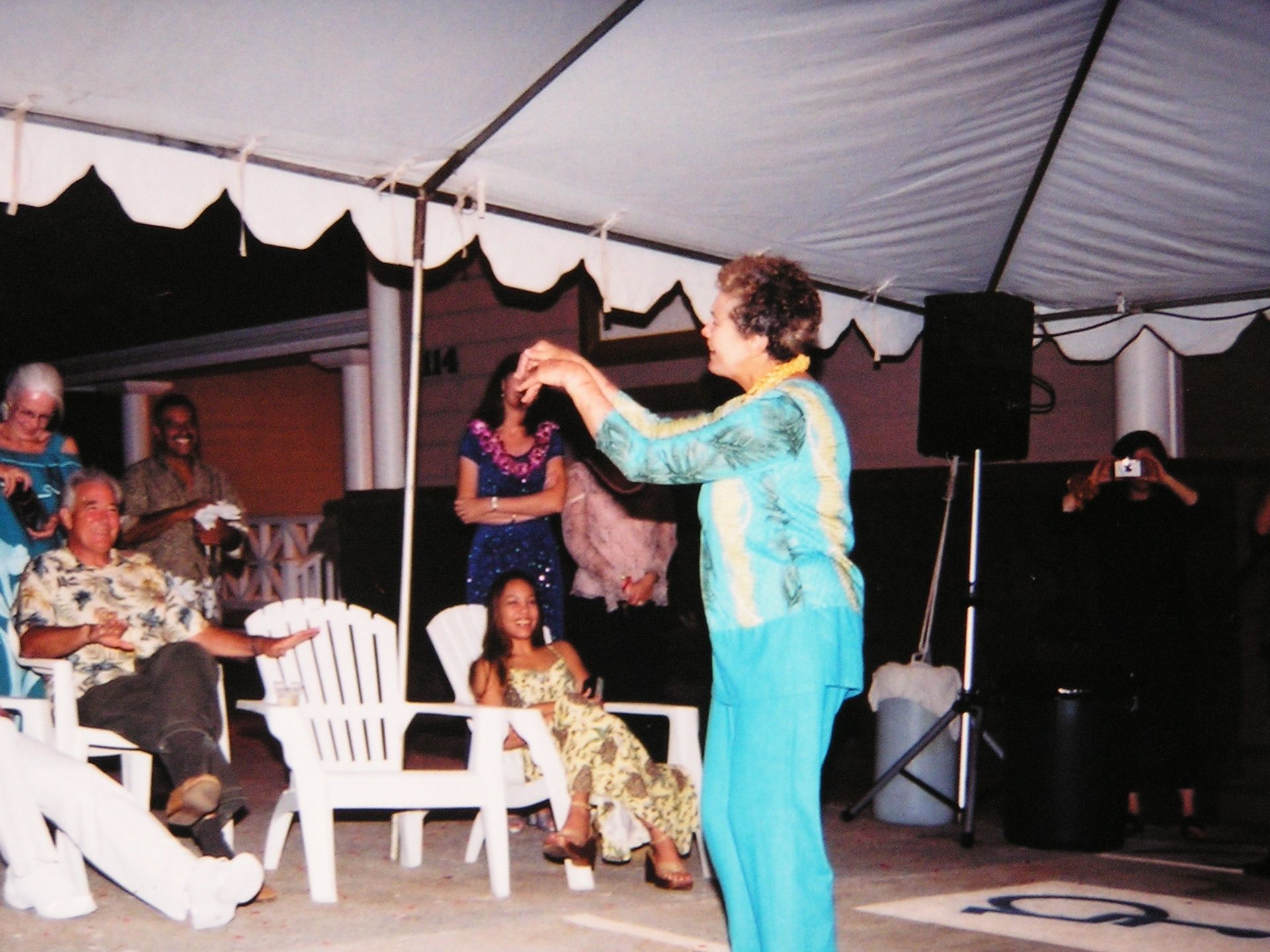 Woman dancing under a tent, gesturing with arms. People watch from chairs. Evening setting.