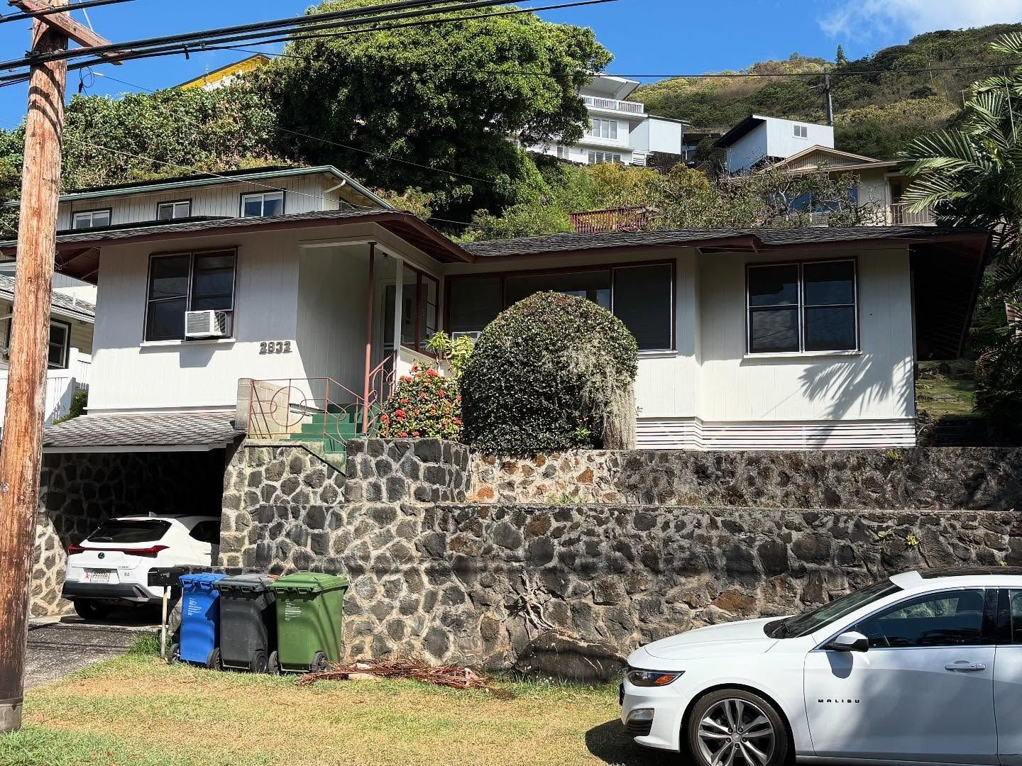 Cottage-style home with carport, stone wall, and parked cars. Trees in the background. Blue, green, and black trash bins in front.