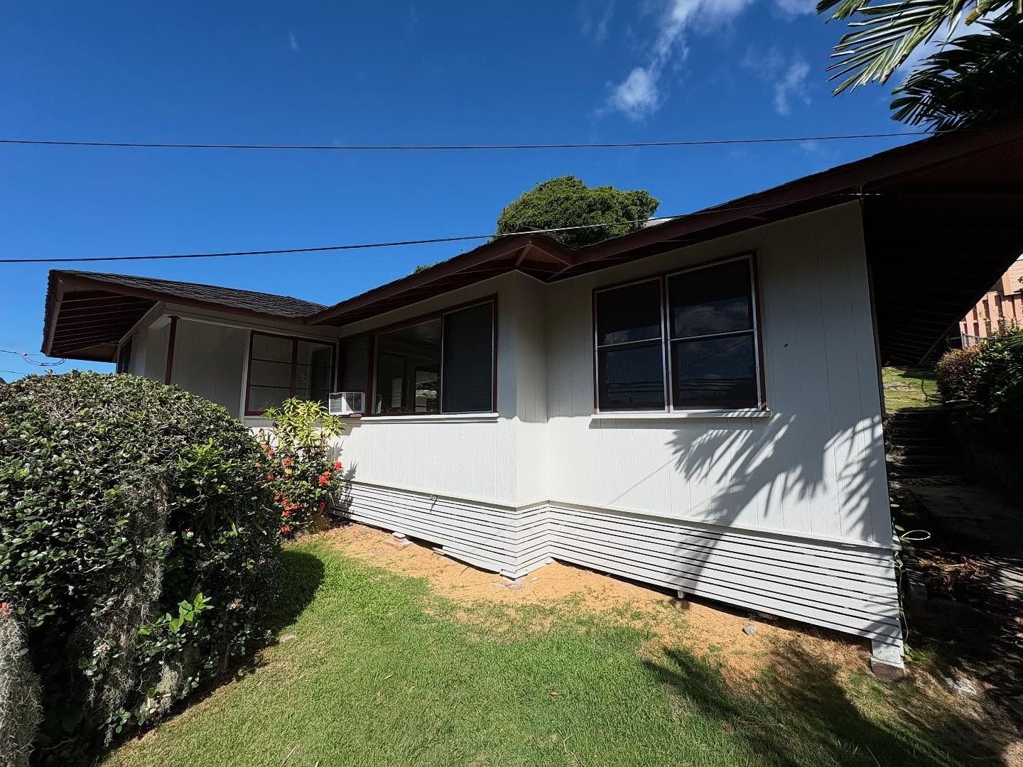 White house with dark brown trim, windows, and carport; green grass and bushes, sunny blue sky.