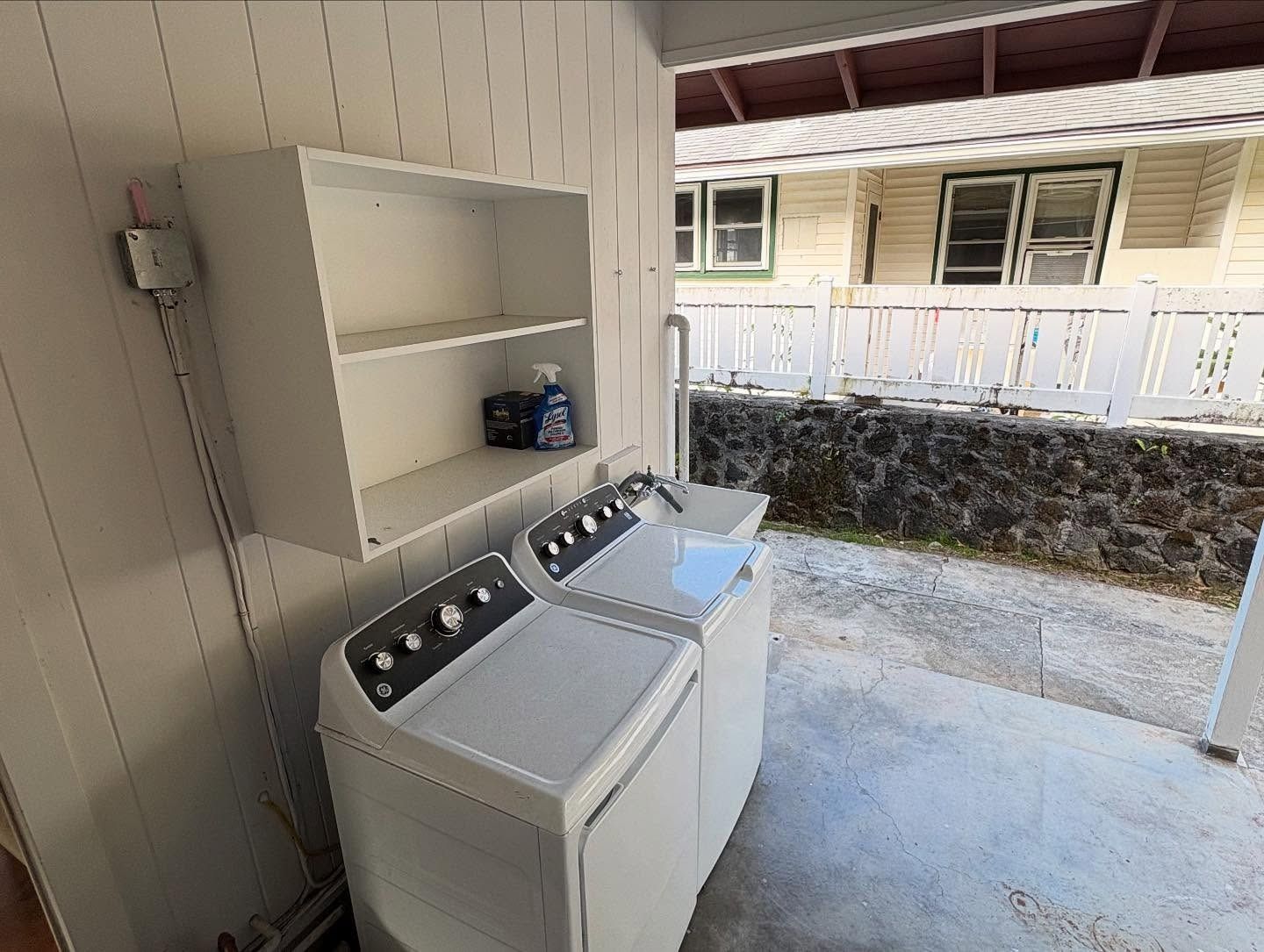 Washing machine and dryer set under a shelf with cleaning supplies. Outdoor laundry area.